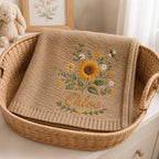May include: A tan knitted blanket featuring an embroidered design of a large sunflower, smaller white and yellow flowers, green leaves, and a small bee. The name Chloe is embroidered in cursive script below the floral arrangement.