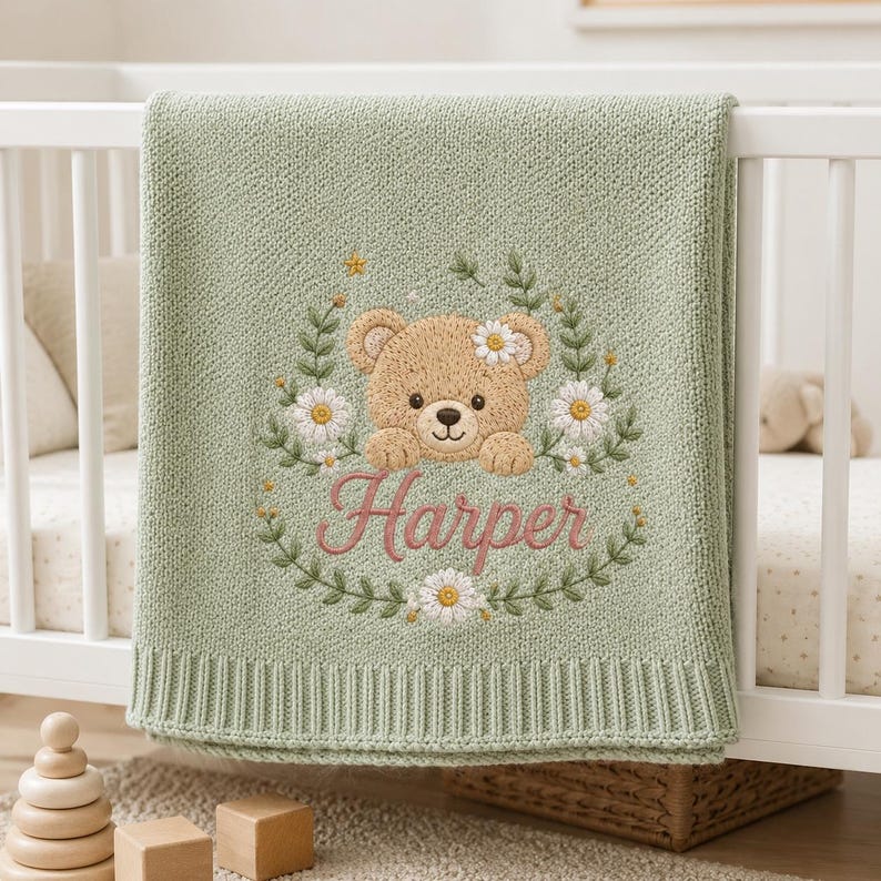 May include: A sage green knitted baby blanket featuring an embroidered design of a tan teddy bear surrounded by a floral wreath of white daisies and green leaves. The name Harper is embroidered in pink script lettering below the bear.