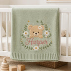 May include: A sage green knitted baby blanket featuring an embroidered design of a tan teddy bear surrounded by a floral wreath of white daisies and green leaves. The name Harper is embroidered in pink script lettering below the bear.
