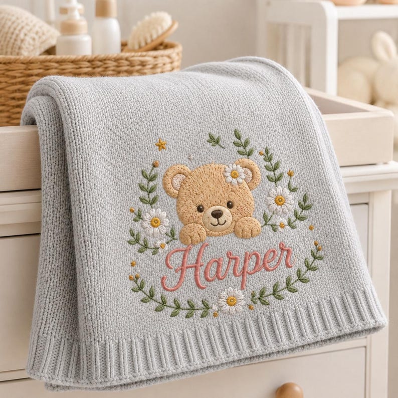 May include: A light gray knitted baby blanket featuring an embroidered design of a tan teddy bear surrounded by green leaves and white daisies. The name Harper is embroidered in pink cursive script below the bear, centered on the fabric.