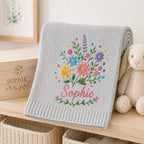 May include: A light gray knit blanket featuring a textured chevron pattern and a central embroidered bouquet of colorful wildflowers in shades of pink, yellow, blue, and purple, with the name Sophie stitched in pink cursive script below th