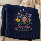 May include: A navy blue knitted blanket featuring a textured surface and a ribbed border. The center displays an embroidered bouquet of colorful flowers in shades of pink, yellow, blue, and purple, with the name Sophie stitched in pink scr