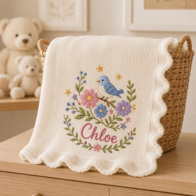 May include: A cream-colored knit blanket with a scalloped edge, featuring an embroidered design of a blue bird perched on a branch surrounded by pink, purple, and blue flowers, green leaves, and yellow stars, with the name Chloe in pink sc
