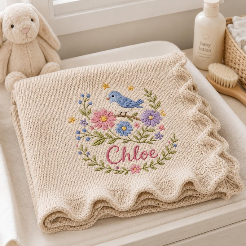 May include: A beige knit baby blanket with a scalloped edge, featuring an embroidered design of a blue bird perched on a branch surrounded by pink, purple, and blue flowers, green leaves, and yellow stars, with the name Chloe in pink scrip