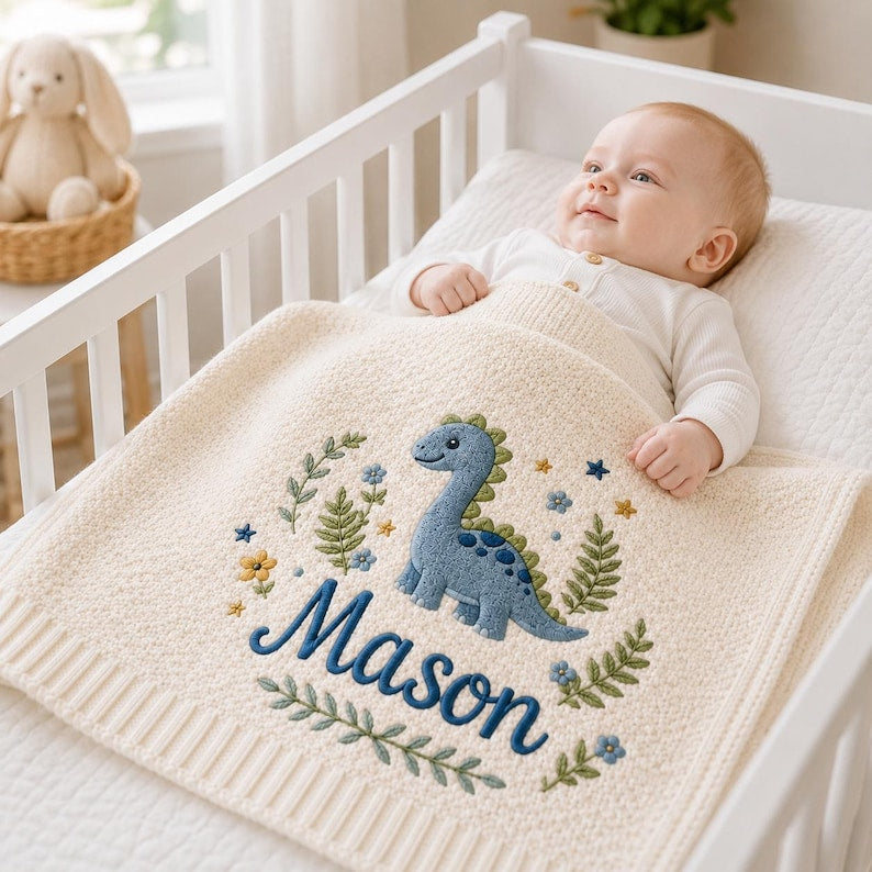 May include: A cream-colored knit baby blanket featuring an embroidered blue dinosaur, green foliage, and small flowers. The name Mason is stitched in dark blue cursive script across the center of the blanket, which has a ribbed border edge