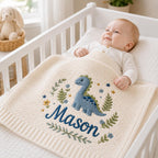 May include: A cream-colored knit baby blanket featuring an embroidered blue dinosaur, green foliage, and small flowers. The name Mason is stitched in dark blue cursive script across the center of the blanket, which has a ribbed border edge