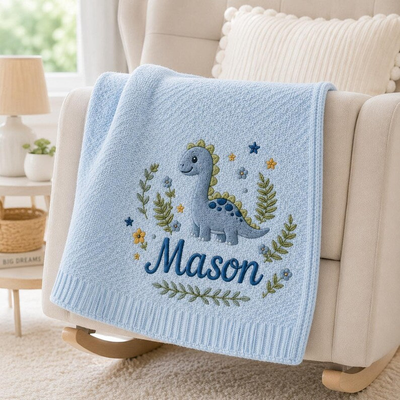 May include: A light blue knit baby blanket featuring an embroidered design of a blue dinosaur surrounded by green foliage, small yellow and blue flowers, and stars. The name Mason is embroidered in dark blue script lettering below the dino