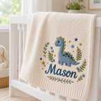 May include: A cream-colored knit blanket featuring an embroidered design of a blue dinosaur surrounded by green foliage, small blue and yellow flowers, and stars. The name Mason is embroidered in dark blue script lettering below the dinosa