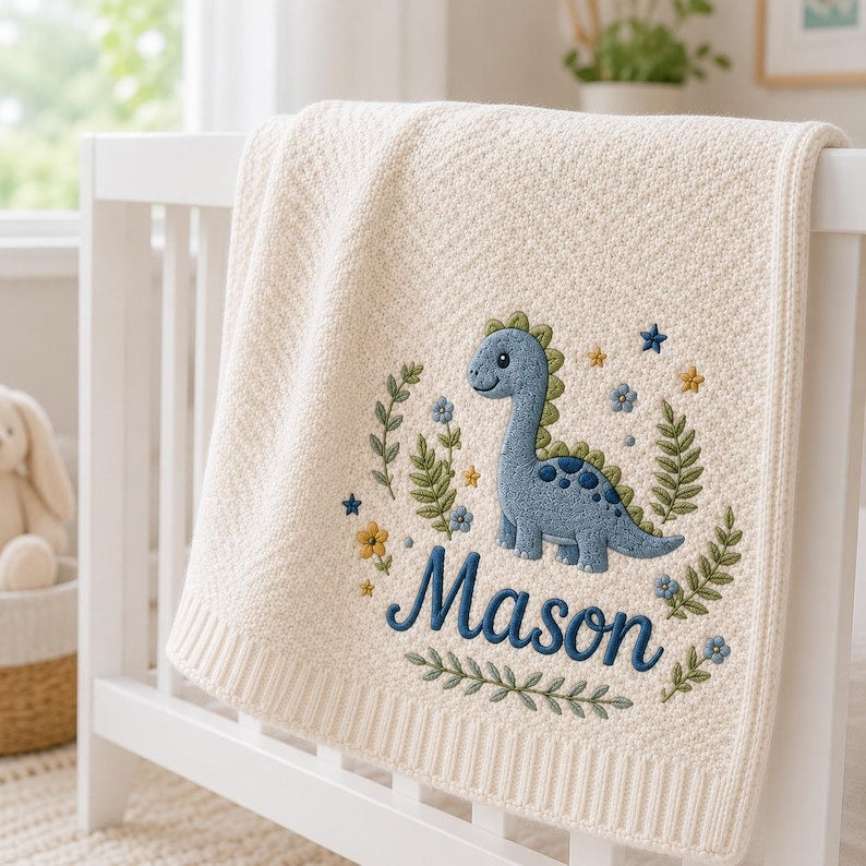May include: A cream-colored knit blanket featuring an embroidered design of a blue dinosaur surrounded by green foliage, small blue and yellow flowers, and stars. The name Mason is embroidered in dark blue script lettering below the dinosa