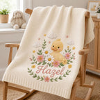 May include: A cream-colored knit baby blanket featuring an embroidered design of a yellow duckling surrounded by white and pink flowers, green leaves, small stars, and clouds. The name Hazel is embroidered in pink script below the duckling