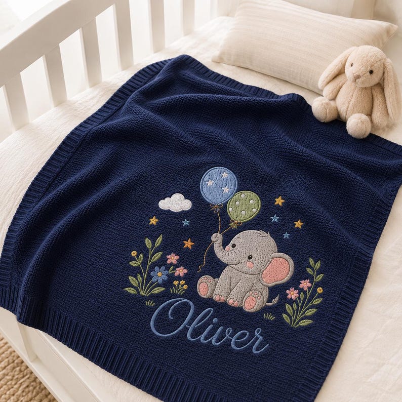 May include: A navy blue knitted baby blanket featuring an embroidered design of a gray elephant holding a blue balloon with white stars and a green polka-dot balloon. The blanket includes floral accents, small stars, a cloud, and the name Oliver.
