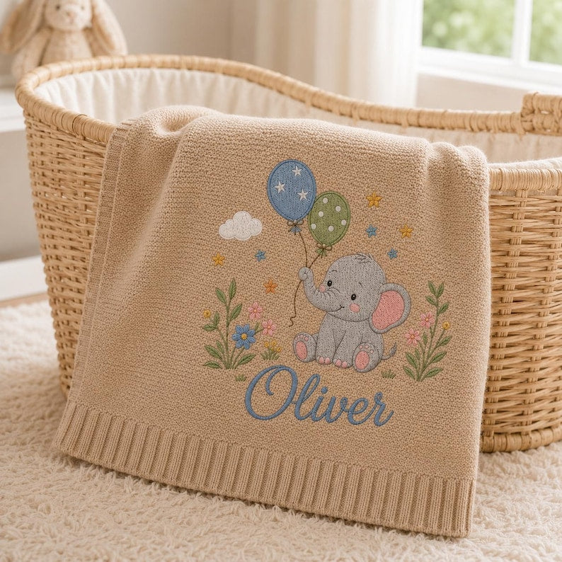 May include: A beige knitted baby blanket featuring an embroidered gray elephant holding two balloons, one blue with white stars and one green with white dots. The design includes small flowers, a white cloud, and the name Oliver in blue script text.