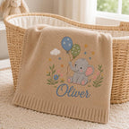 May include: A beige knitted baby blanket featuring an embroidered gray elephant holding two balloons, one blue with white stars and one green with white dots. The design includes small flowers, a white cloud, and the name Oliver in blue script text.