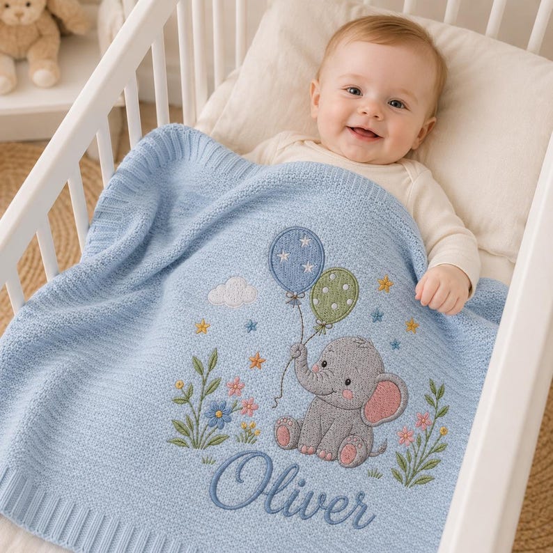 May include: A light blue knitted baby blanket featuring an embroidered gray elephant holding a blue balloon with white stars and a green polka-dot balloon. The design includes colorful flowers, small stars, and the name Oliver in blue script.