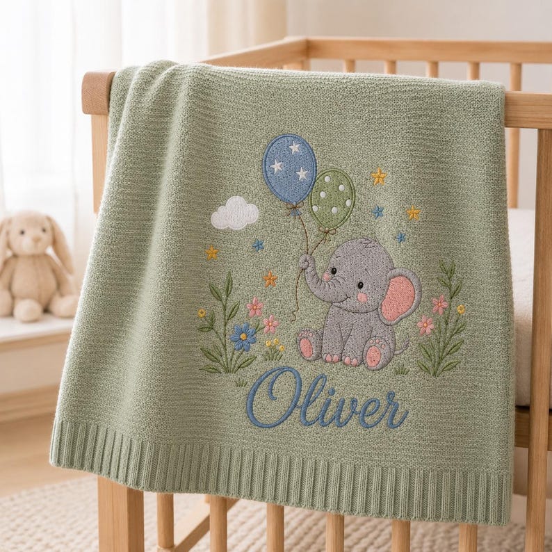 May include: A sage green knitted baby blanket featuring an embroidered gray elephant holding two balloons, one blue with stars and one green with dots. The design includes small flowers, a white cloud, and the name Oliver embroidered in blue script.