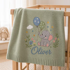 May include: A sage green knitted baby blanket featuring an embroidered gray elephant holding two balloons, one blue with stars and one green with dots. The design includes small flowers, a white cloud, and the name Oliver embroidered in blue script.