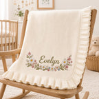 May include: A cream-colored knit baby blanket featuring a ruffled edge and an embroidered floral garland design. The name Evelyn is stitched in green cursive script in the center of the flowers, which include white daisies and pink blossoms.