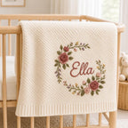 May include: A cream-colored knit baby blanket featuring a diagonal ribbed texture and a central embroidered floral wreath in shades of pink, green, and yellow. The name Ella is embroidered in a cursive script in the center of the floral wreath.