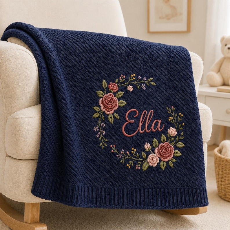 May include: A navy blue knitted blanket featuring a diagonal ribbed texture and a central embroidered floral wreath in shades of pink and green. The name Ella is embroidered in a cursive pink script in the center of the floral wreath.