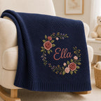 May include: A navy blue knitted blanket featuring a diagonal ribbed texture and a central embroidered floral wreath in shades of pink and green. The name Ella is embroidered in a cursive pink script in the center of the floral wreath.