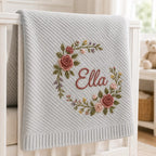 May include: A light gray knitted blanket featuring a diagonal ribbed texture and a central embroidered floral wreath in shades of pink, green, and yellow. The name Ella is embroidered in a cursive script in the center of the floral wreath.