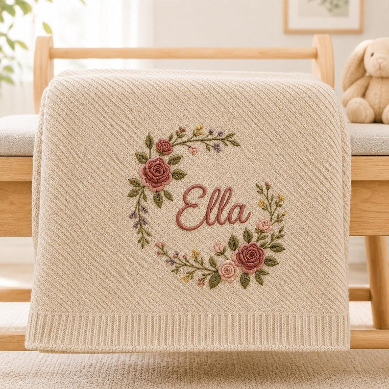 May include: A cream-colored knit blanket featuring a diagonal ribbed texture and a central embroidered floral wreath. The wreath surrounds the name "Ella" stitched in a dark pink cursive font, with roses in shades of pink and green leaves.