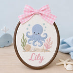 May include: An oval embroidery hoop featuring a light blue octopus, green seaweed, pink coral, and seashells stitched on white fabric. The name Lily is embroidered in pink script below the sea life. A pink and white gingham bow sits on top
