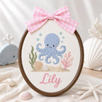 May include: An oval embroidery hoop featuring a blue octopus, green seaweed, pink coral, and seashells stitched on white fabric. The name Lily is embroidered in pink script below the sea life. A pink and white gingham bow sits at the top.