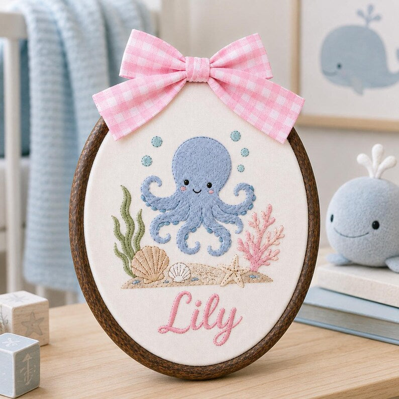 May include: An oval embroidery hoop featuring a blue octopus, green seaweed, pink coral, and seashells stitched on white fabric. The name Lily is embroidered in pink script below the sea life. A pink and white gingham bow is attached at th
