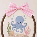 May include: A circular wooden embroidery hoop containing a fabric panel with a hand-stitched blue octopus, green seaweed, pink coral, and seashells. A pink and white gingham fabric bow is attached to the top of the dark brown wooden frame.