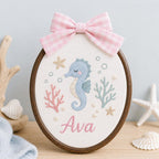 May include: An oval embroidery hoop featuring a light blue seahorse, pink coral, and green seaweed stitched on white fabric. The name Ava is embroidered in pink script below the seahorse. A pink and white gingham bow is attached to the top