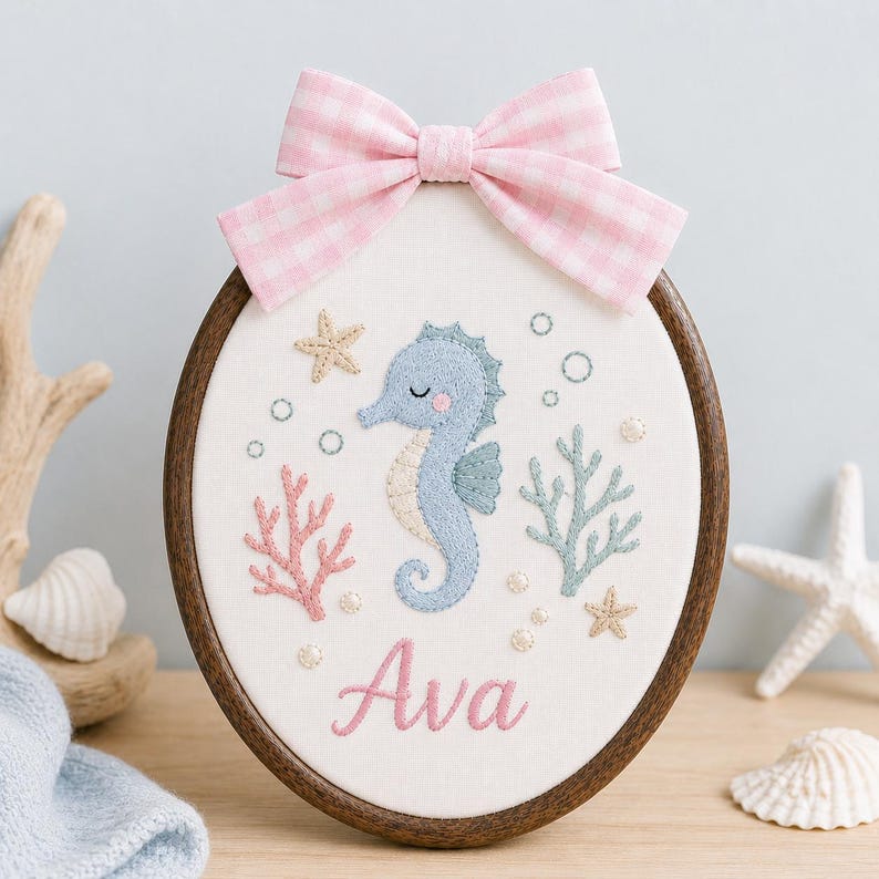 May include: An oval embroidery hoop featuring a light blue seahorse, pink coral, and green seaweed stitched on white fabric. The name Ava is embroidered in pink script below the seahorse. A pink and white gingham bow is attached to the top