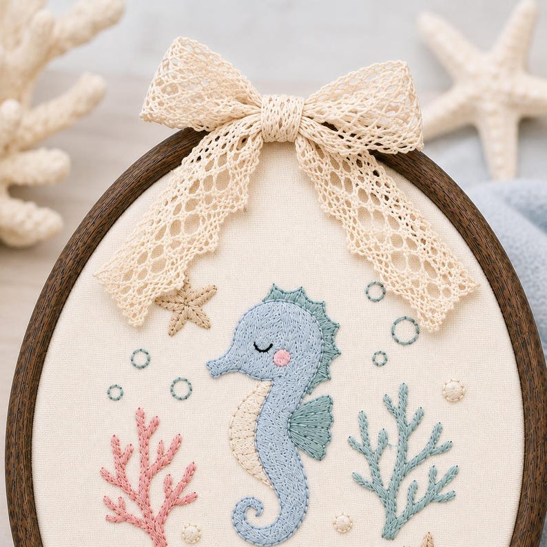 May include: An oval embroidery hoop containing a cream-colored fabric panel with a stitched light blue seahorse, pink and teal coral, and small bubbles. A beige lace bow is attached to the top of the dark wood frame, which is circular in s