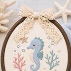 May include: An oval embroidery hoop containing a cream-colored fabric panel with a stitched light blue seahorse, pink and teal coral, and small bubbles. A beige lace bow is attached to the top of the dark wood frame, which is circular in s