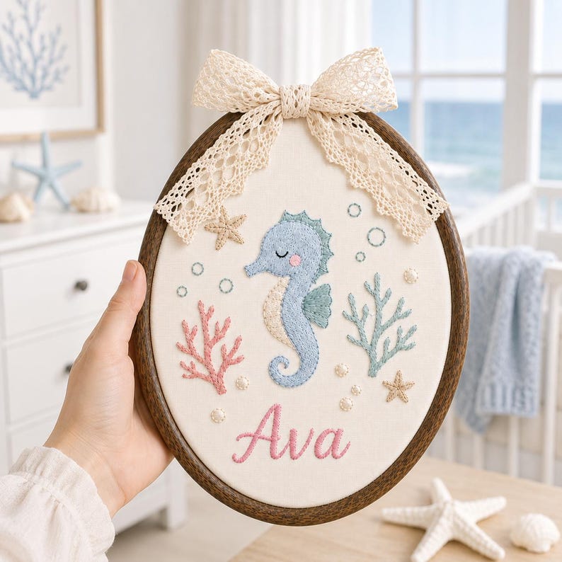 May include: An oval wooden embroidery hoop containing a cream-colored fabric panel with a stitched seahorse, pink coral, blue seaweed, and starfish. The name Ava is embroidered in pink script at the bottom, topped with a beige lace bow.