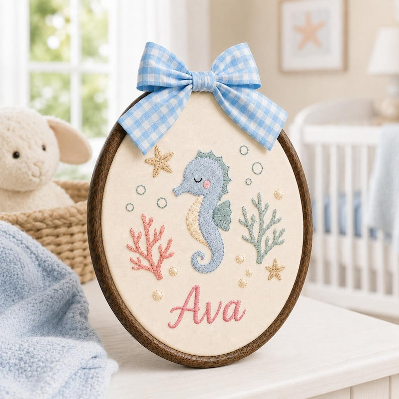 May include: An oval embroidery hoop wall hanging featuring a light blue seahorse, pink coral, green seaweed, and starfish on cream fabric. The name Ava is embroidered in pink script at the bottom, and a blue and white gingham bow sits on t