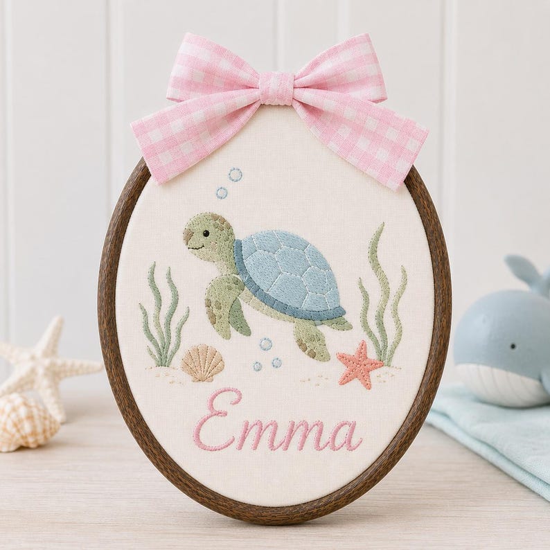 May include: An oval embroidery hoop featuring a light green sea turtle, a small seashell, a pink starfish, and green seaweed stitched on off-white fabric. The name Emma is embroidered in pink script below the turtle, topped with a pink gin