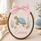 May include: An oval embroidery hoop featuring a light green sea turtle with a blue shell, surrounded by embroidered seaweed, a seashell, a starfish, and blue bubbles. The name Emma is stitched in pink script below, topped with a pink gingh