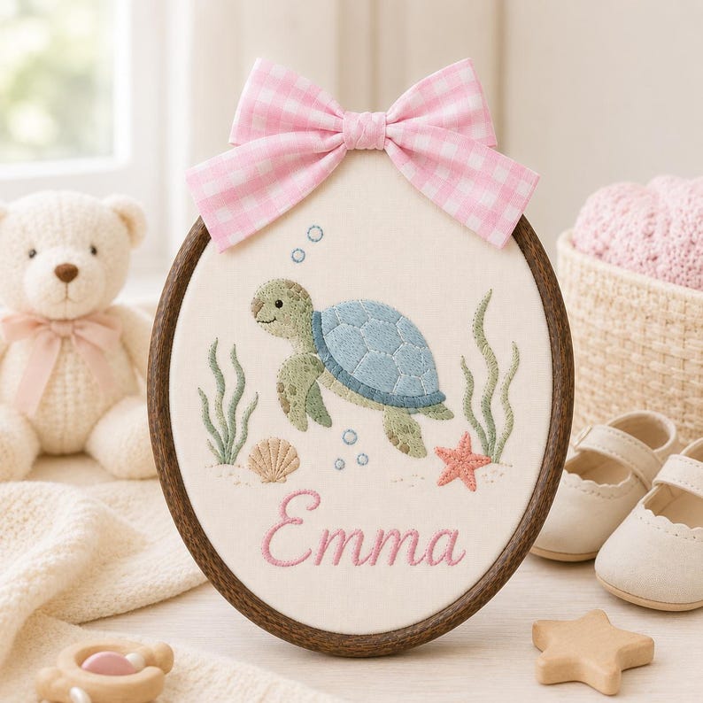 May include: An oval embroidery hoop featuring a light blue and green sea turtle, a pink starfish, a seashell, and green seaweed on a cream fabric background. The name Emma is embroidered in pink script below the turtle, topped with a pink 