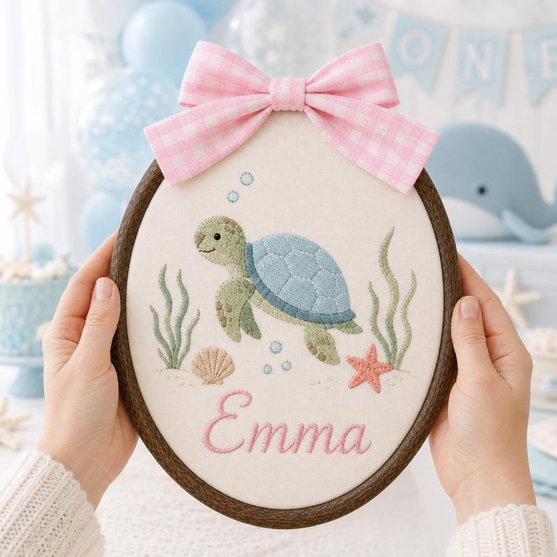 May include: An oval embroidery hoop featuring a light-colored fabric with a stitched sea turtle, seaweed, a seashell, and a starfish. The name Emma is embroidered in pink script below the turtle. A pink and white gingham bow is attached at