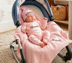 May include: A light pink baby outfit set consisting of a long-sleeved onesie, a knotted beanie, and a knit blanket. The beanie and onesie feature the name Alexander in black embroidery, while the blanket displays the name Alexander in whit