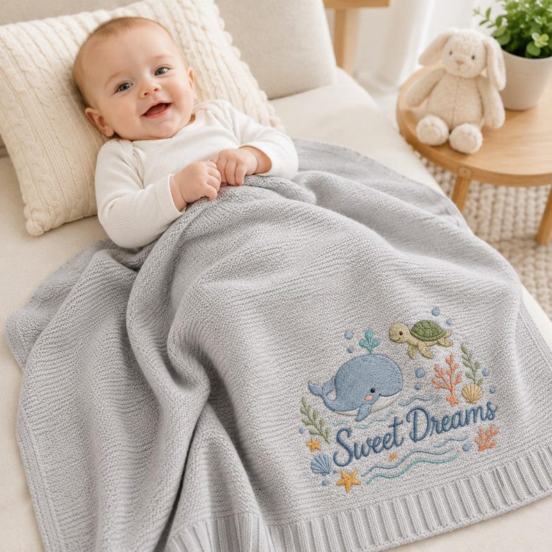 May include: A light gray knitted baby blanket featuring an embroidered design of a blue whale, a green sea turtle, colorful coral, seashells, and starfish. The text "Sweet Dreams" is embroidered in blue script lettering on the blanket surface.