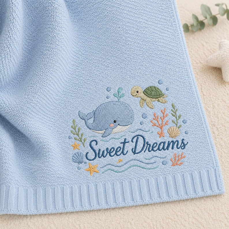 May include: A light blue knit blanket featuring an embroidered design of a whale, a sea turtle, coral, seashells, and starfish. The text Sweet Dreams is embroidered in dark blue script below the whale and turtle, surrounded by ocean motifs.