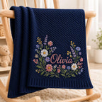 May include: A navy blue knitted blanket featuring a diagonal ribbed texture and an embroidered floral wreath design surrounding the name Olivia in pink script. The blanket is draped over a wooden chair, showcasing detailed colorful wildflo