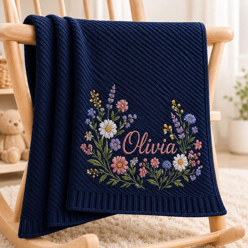 May include: A navy blue knitted blanket featuring a diagonal ribbed texture and an embroidered floral wreath design surrounding the name Olivia in pink script. The blanket is draped over a wooden chair, showcasing detailed colorful wildflo