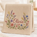 May include: A beige knit blanket featuring a diagonal ribbed texture and a wide ribbed border. The center displays an embroidered floral wreath in shades of pink, blue, yellow, and green, with the name Olivia stitched in a dark pink script