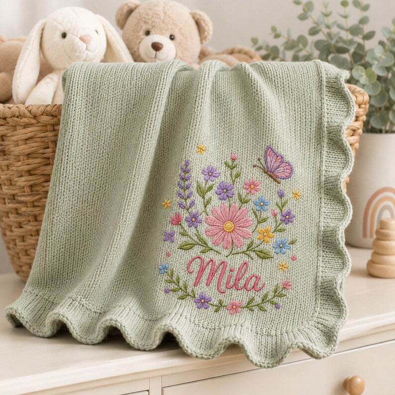 May include: A sage green knitted baby blanket with a scalloped edge, featuring an embroidered design of colorful wildflowers, green leaves, and a purple butterfly. The name Mila is embroidered in pink cursive script below the floral arrang