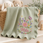 May include: A sage green knitted baby blanket with a scalloped edge, featuring an embroidered design of colorful wildflowers, green leaves, and a purple butterfly. The name Mila is embroidered in pink cursive script below the floral arrang