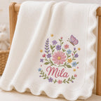 May include: A white knitted baby blanket with a scalloped edge, featuring an embroidered design of colorful flowers, green leaves, and a purple butterfly, with the name Mila stitched in pink cursive script below the floral arrangement.