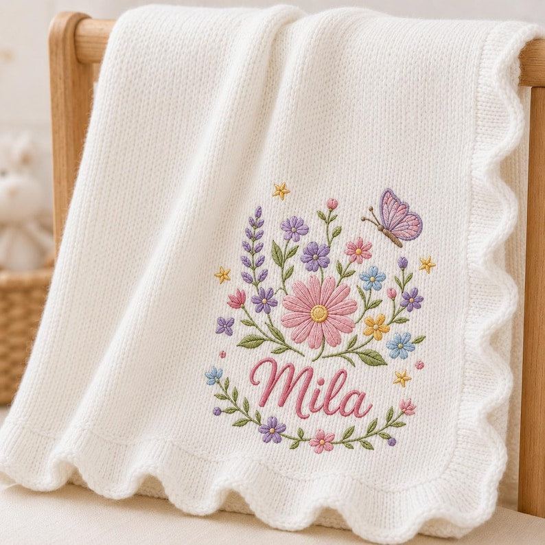 May include: A white knitted baby blanket with a scalloped edge, featuring an embroidered design of colorful flowers, green leaves, and a purple butterfly, with the name Mila stitched in pink cursive script below the floral arrangement.